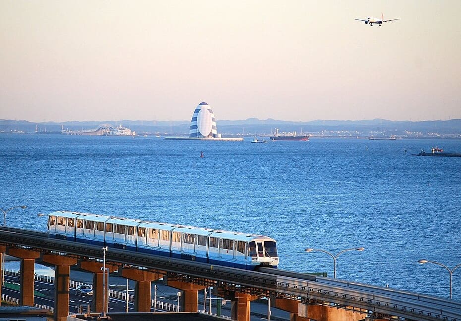 A monorail train in Tokyo near water and ships. An airplane is flying above it.